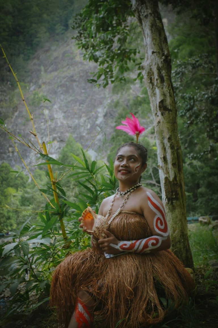 Woman in traditional tribal attire with body paint, seated in a lush forest setting.